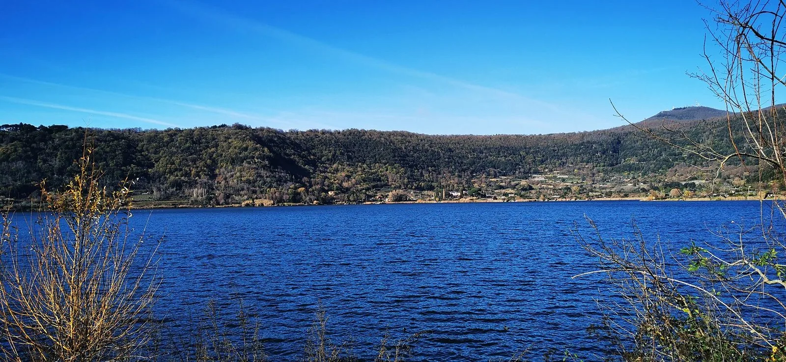 Il lago di Nemi sotto un cielo azzurro, con i pendii del cratere coperti di boschi dai toni autunnali e rami spogli in primo piano